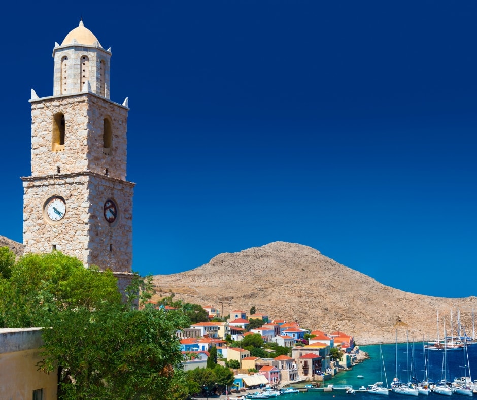 Kamienna wieża zegarowa w miasteczku Chalki, zielone drzewa, niebieskie bezchmurne niebo, wyspa Chalki, Rodos, Grecja